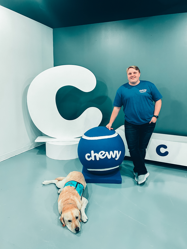 Casey Lavender, a CVM student, stands next to a large tennis ball with the word "Chewy" on it. A mid sized dog is next to him.